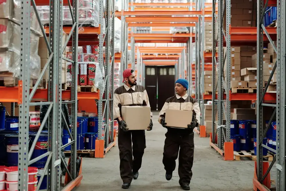 Two warehouse workers carrying boxes through a logistics facility.